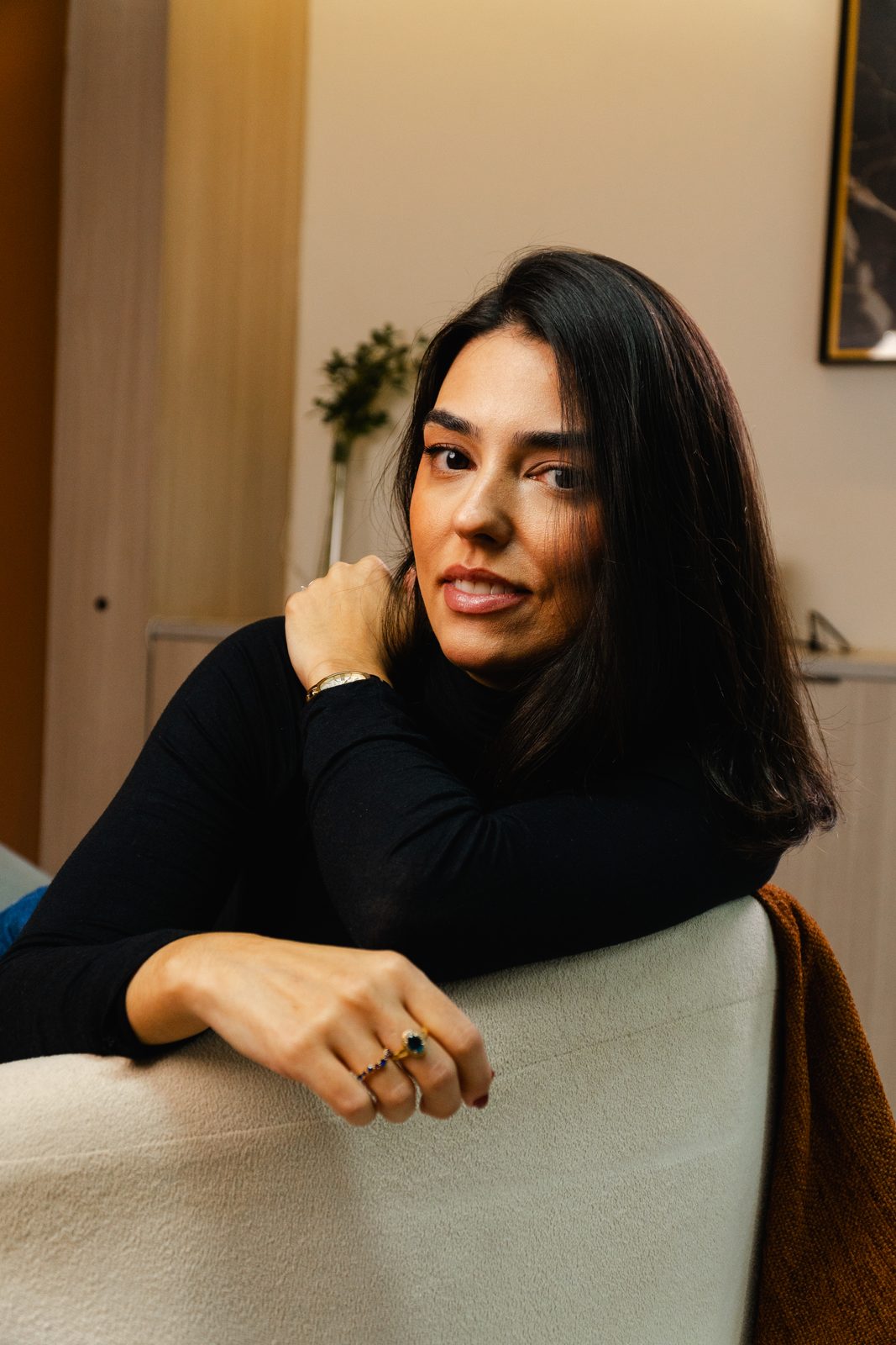 Thaís Dal Piaz, de blusa preta, apoiada no encosto de uma poltrona, olhando para a câmera