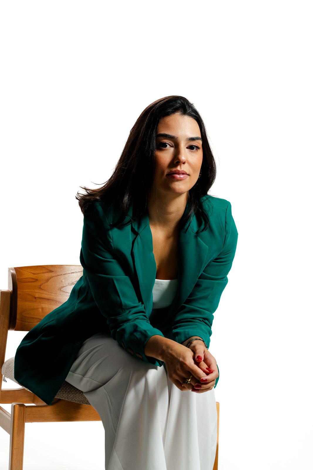 Thaís Dal Piaz sentada na beira de uma cadeira, de blazer verde, olhando para a câmera