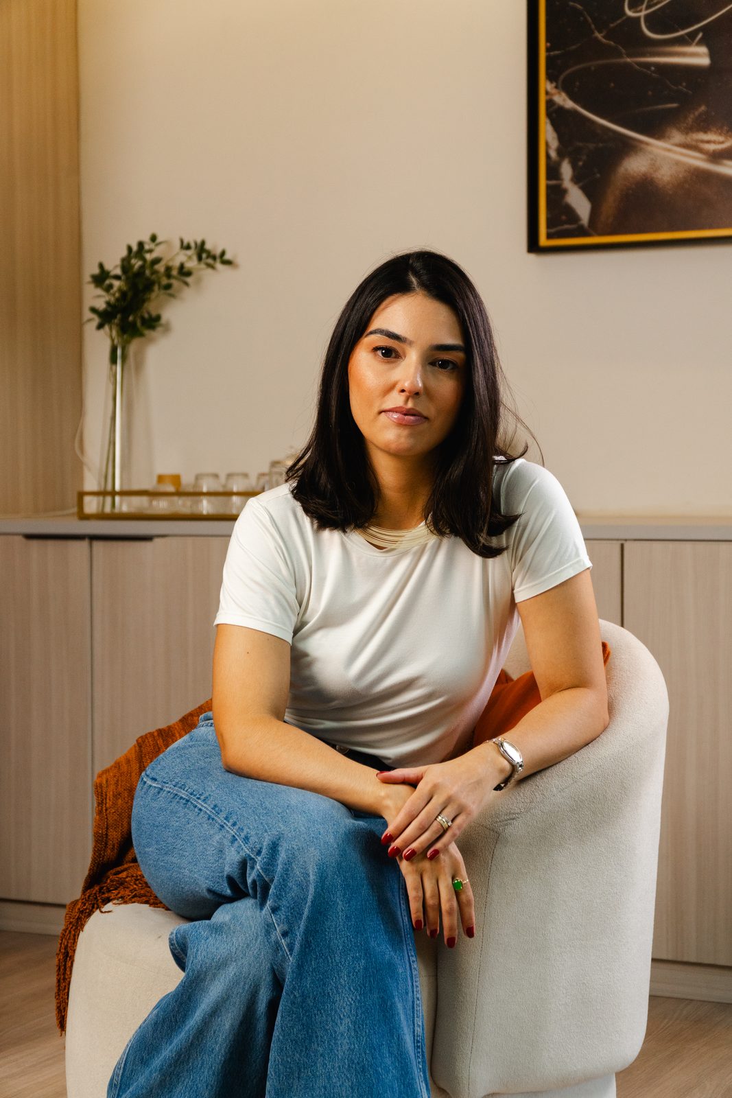 Thaís Dal Piaz sentada à mesa de trabalho, de camiseta branca, com um quadro ao fundo
